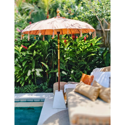 A sunlit terracotta Balinese parasol shading a poolside lounge bed with neutral-toned cushions