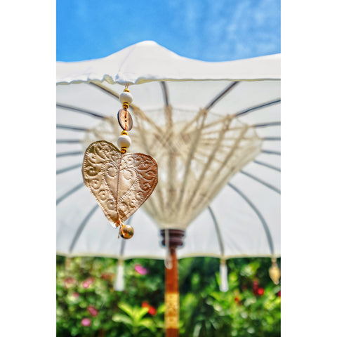 Golden heart ornament and tassel hanging from the edge of the white parasol, with the canopy and floral garden in soft focus.