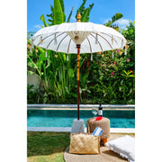 Front-facing image of the white parasol fully open by a villa pool, showing intricate internal weave and decorative top finial.